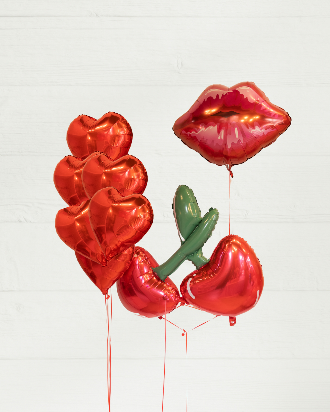 A balloon bouquet featuring three red heart-shaped foil balloons, an oversized cherry-shaped balloon, and a red lips-shaped balloon, arranged together.