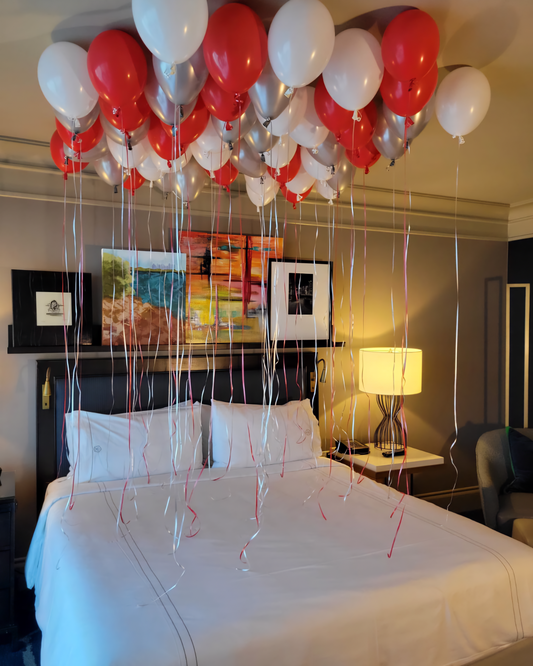 A bedroom decorated with a heart-shaped arrangement of red and white balloons, with balloons scattered around the room and tied to the bedposts.