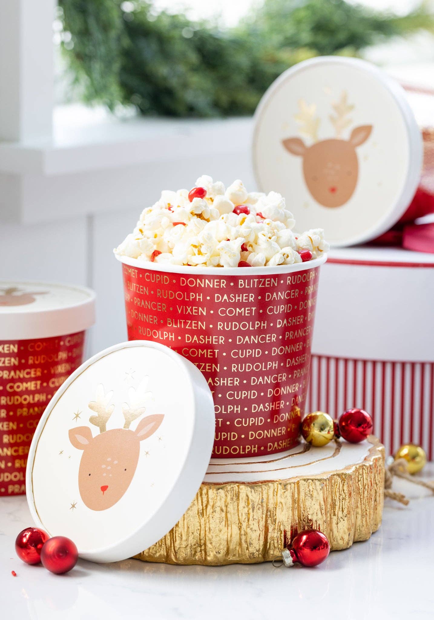 Popcorn in a festive container with reindeer design on a wooden log, surrounded by Christmas decorations.