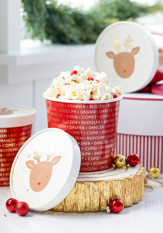 Popcorn in a festive container with reindeer design on a wooden log, surrounded by Christmas decorations.