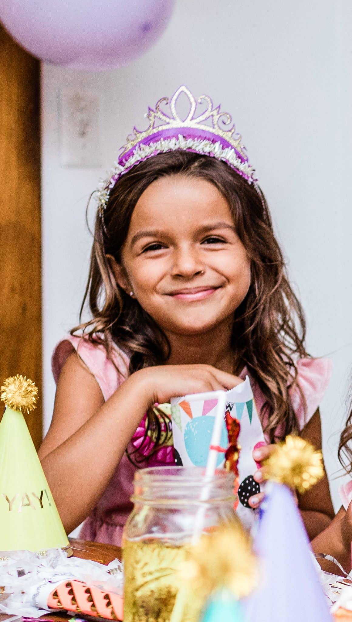 Young girl wearing a purple tiara at a birthday party with balloons and decorations.