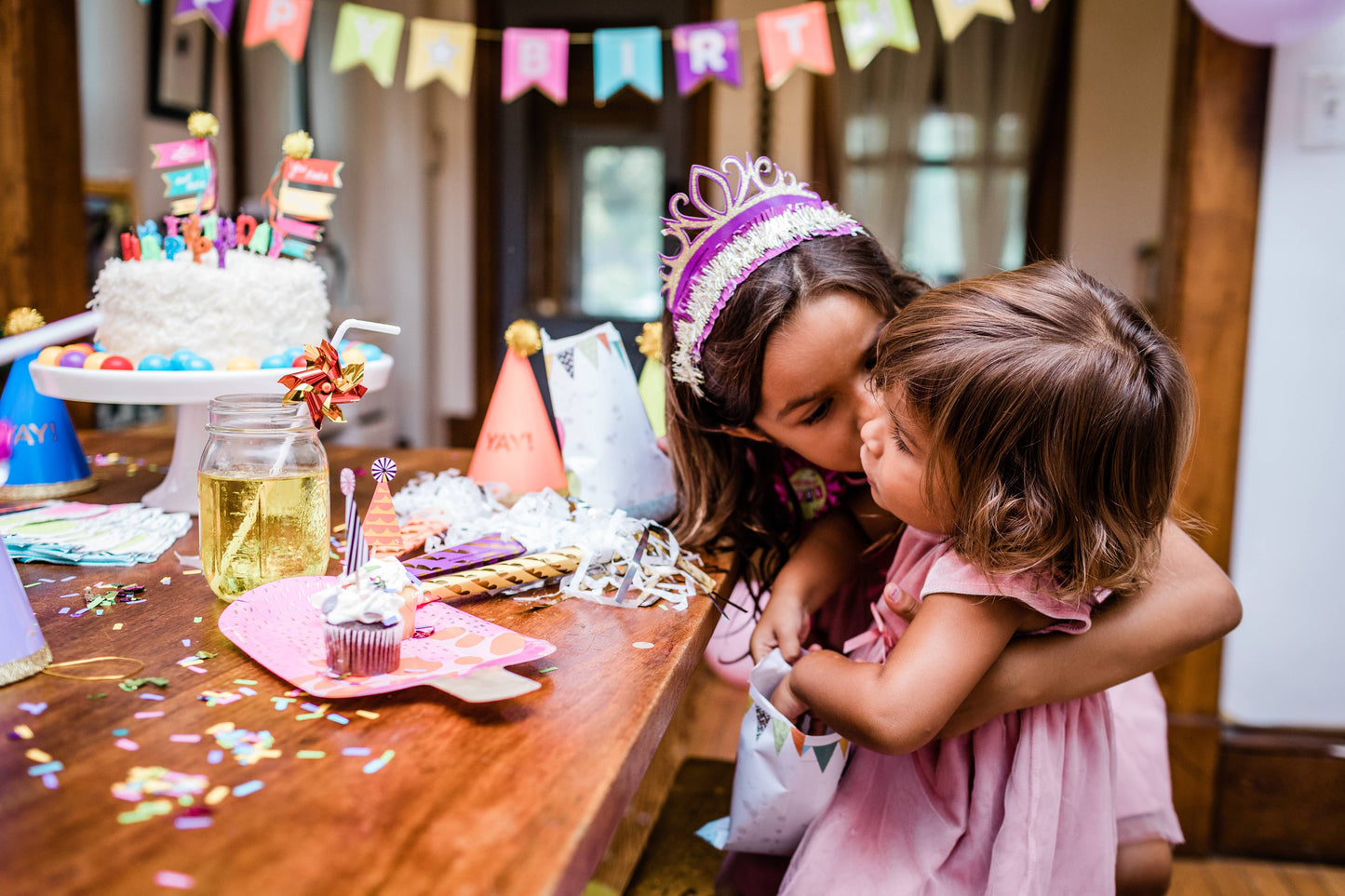 Two young girls hugging at a birthday party with a cake and decorations in the background.